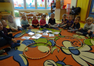 Dzieci siedzą przed rozłożonymi na dywanie obrazkami szczoteczek w różnym kolorze i wielkości oraz dwiema obręczami.